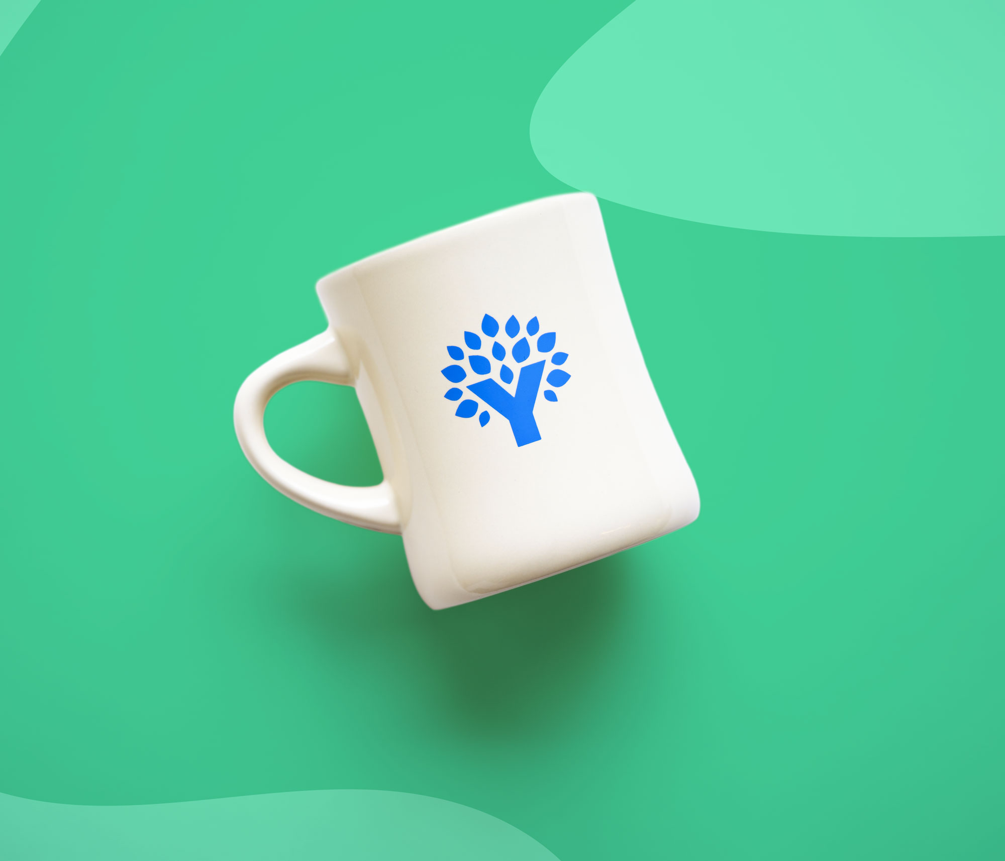 Cream ceramic mug with the YNAB tree logo printed in blue, displayed on a teal background.