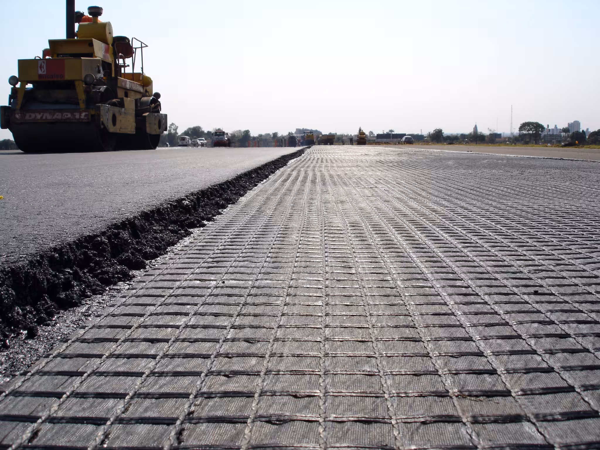 Close-up of road paving with a grid pattern and a large yellow road roller on the left side.