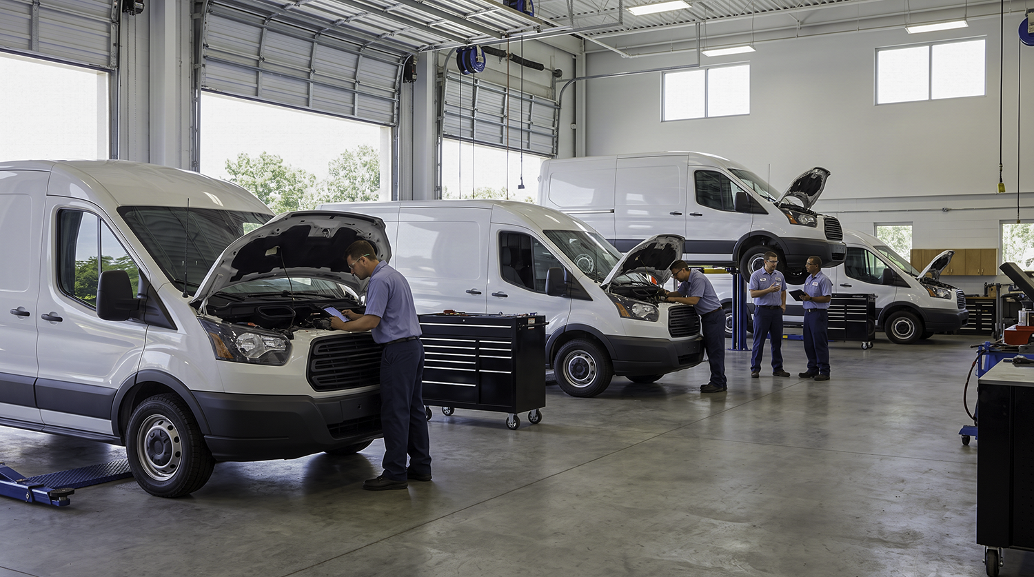 Technicians performing scheduled fleet maintenance on a row of white cargo vans, one using a tablet to read diagnostic codes