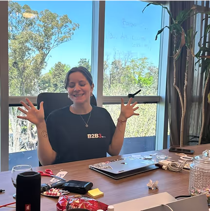 B2B3 team member smiling enthusiastically at her workspace with laptop and office supplies, framed by large windows with greenery views