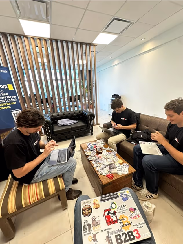 B2B3 team working in modern coworking lounge with wooden dividers, featuring laptops covered in crypto stickers and casual collaboration environment