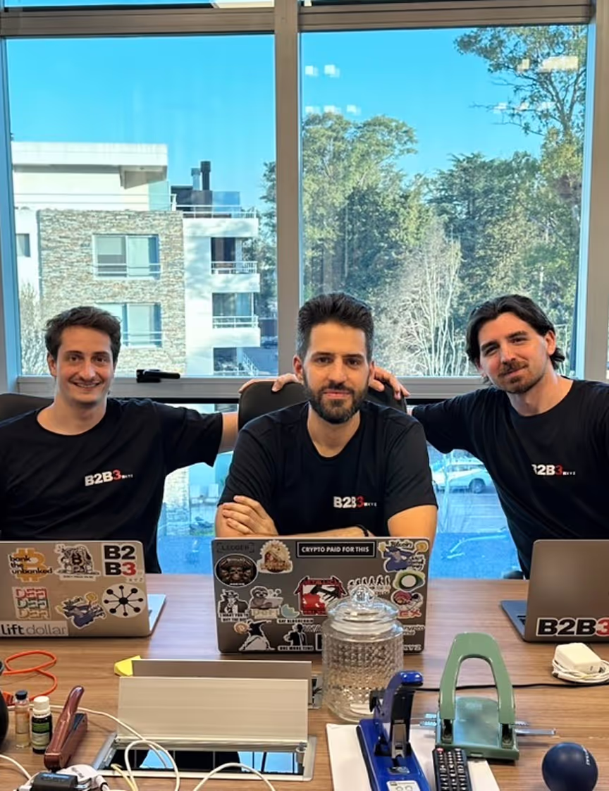 Three B2B3 founders - Mariano Perez, Felipe Lolster, and Matias Soto Quintus - seated at workspace with crypto-decorated laptops against modern office backdrop with panoramic views