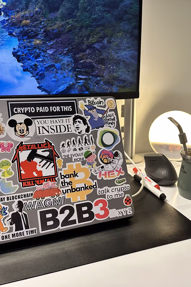 Laptop decorated with crypto-themed stickers featuring Bitcoin wizard, evolution of money illustration, and blockchain references in B2B3's creative workspace