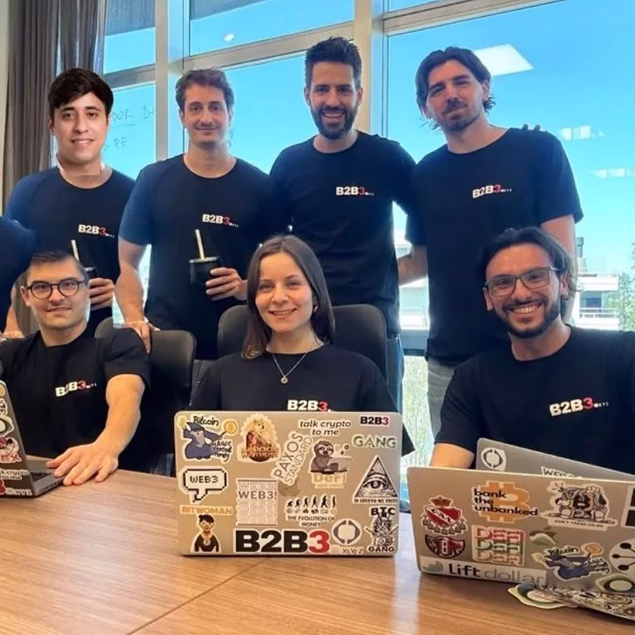 B2B3 team members gathered in modern office space wearing branded shirts, with crypto-themed laptop stickers showcasing the company's Web3 and blockchain expertise