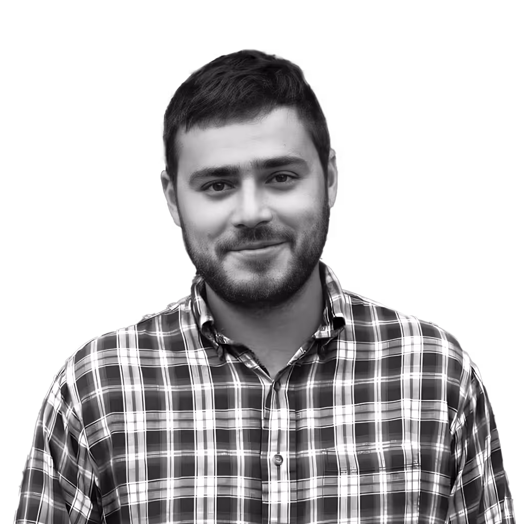 Smiling man with short hair and beard wearing a plaid shirt standing in front of a brick wall.