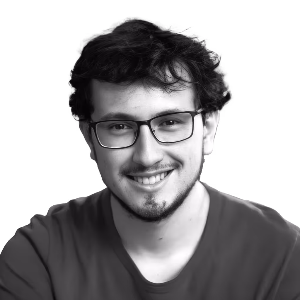 Smiling young man with curly hair, glasses, and a beard wearing a plain shirt.