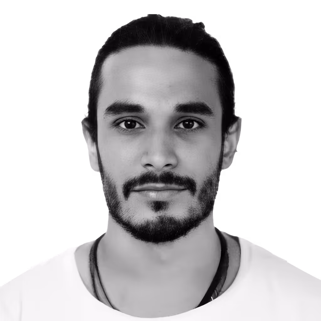 Portrait of a young man with dark hair tied back, a beard, and mustache, wearing a white shirt and necklace, looking directly at the camera.