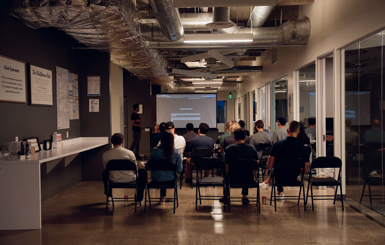 People seated on folding chairs watching a presenter in a modern conference room with exposed ductwork and a projection screen.
