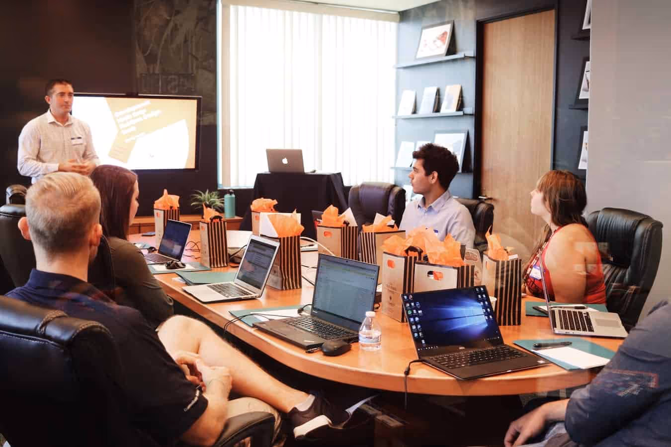 A group of people sitting around a conference table with laptops while a man presents near a screen in a meeting room.