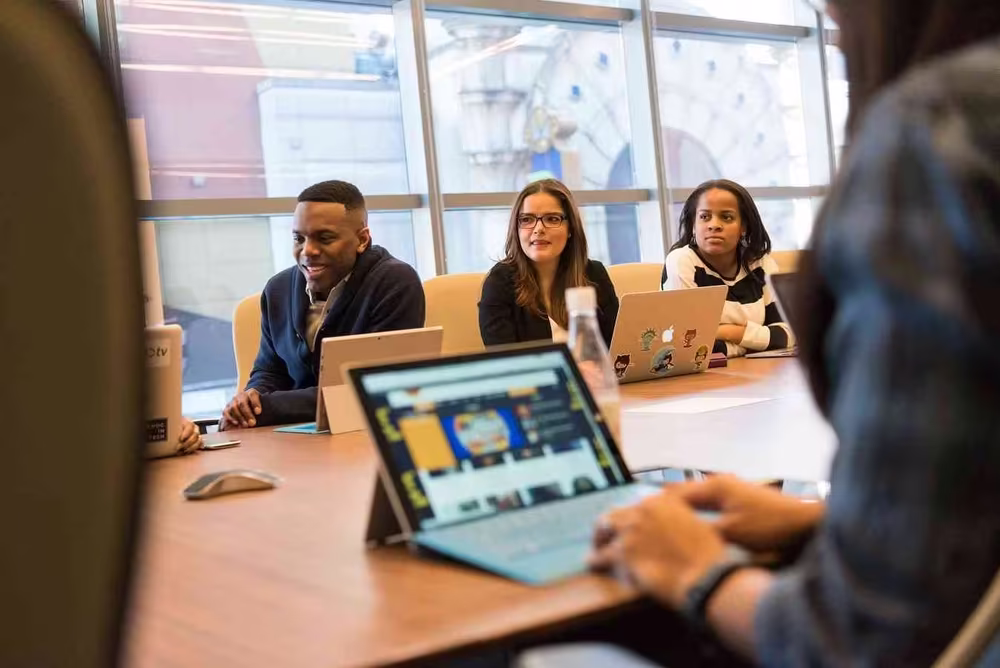 A diverse group of people sitting at a conference table with laptops, engaged in a discussion in a modern office with large windows.