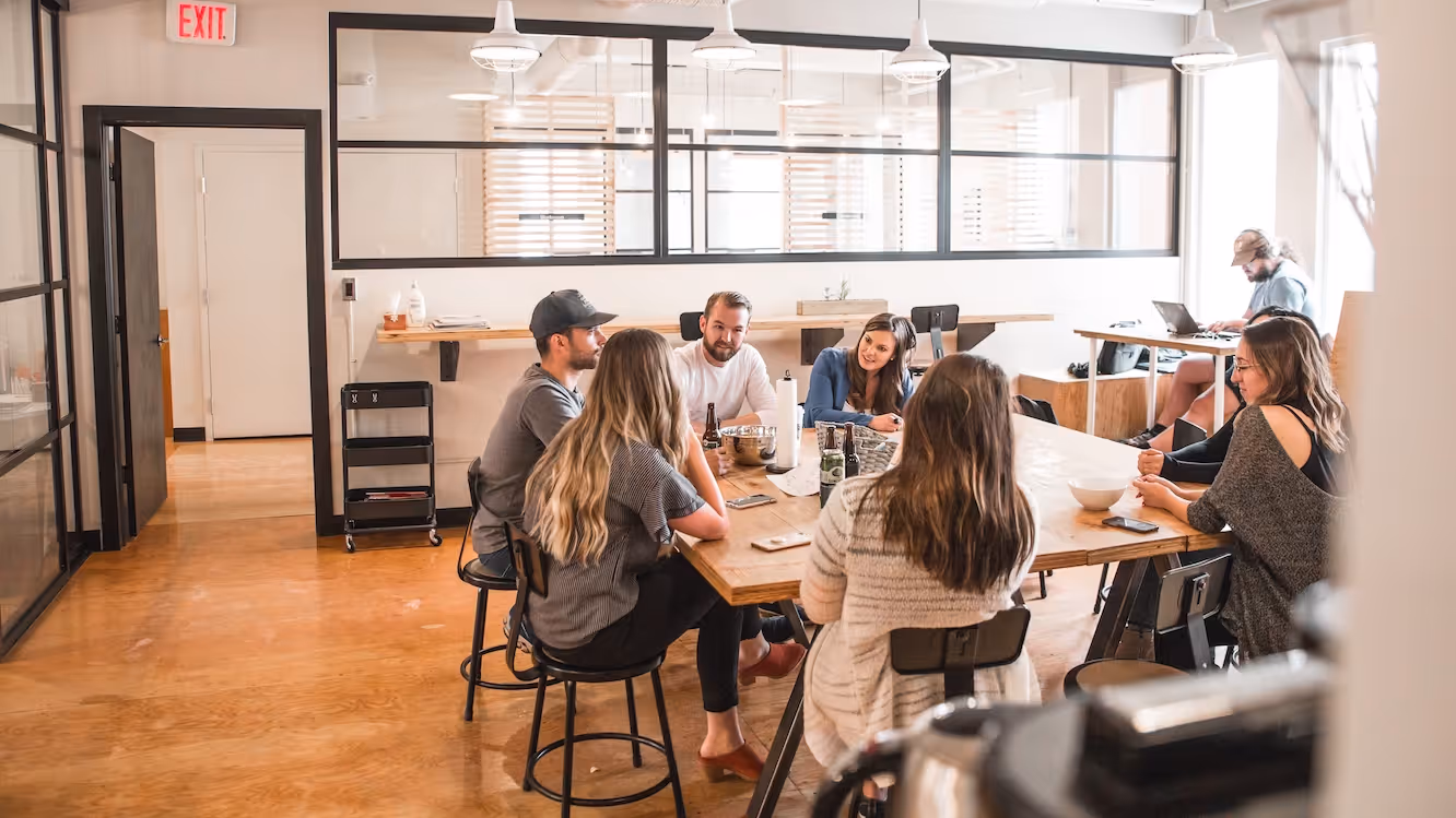 Group of people sitting around a wooden table in a bright office space, engaged in conversation and working.