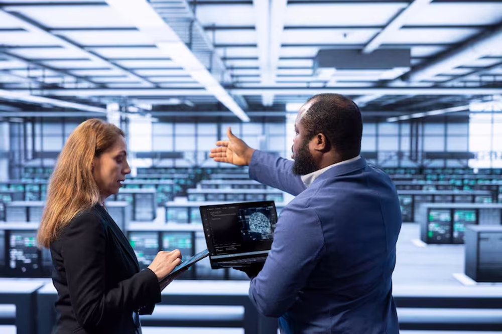 Two data center technicians in business attire discussing while viewing a laptop displaying AI technology in a large server room.