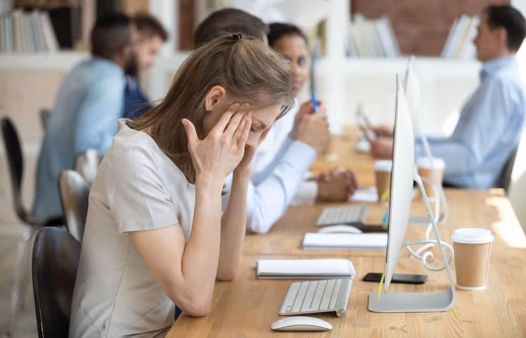 Woman sitting at office desk holding her head in frustration with blurred colleagues working in the background.