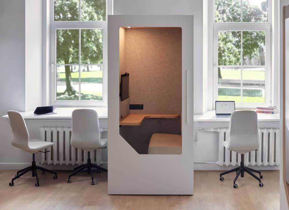 Modern office space with a white phone booth having a built-in desk and chair, surrounded by three grey swivel chairs at window-side desks.
