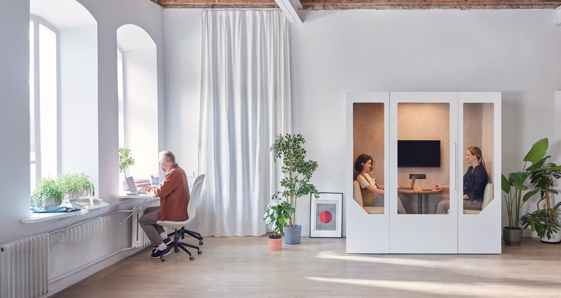 Modern office space with a man working on a laptop by tall windows and two women sitting inside a white phone booth having a conversation.