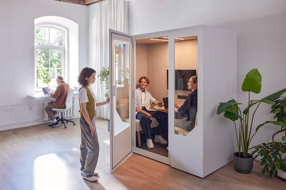 Person taking a phone call inside a soundproof office booth within an open plan workspace