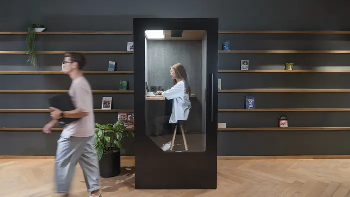 Person_working_inside_a_dark_acoustic_office_pod_with_colleague_walking_past_in_open_office