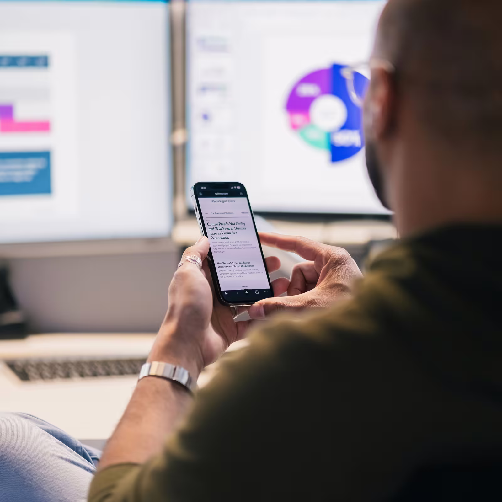 Employee distracted by phone while work screens with charts and data sit unattended in the background, showing divided attention and reduced focus