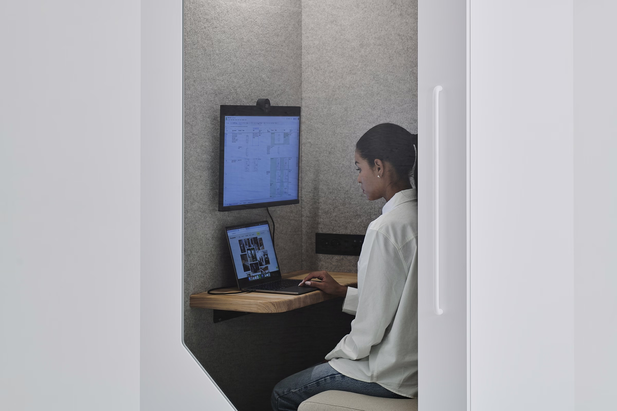A woman working on a laptop inside an office pod with grey felt acoustic lining and a wall-mounted monitor