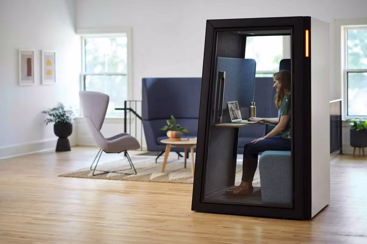Woman working on a laptop inside a black phone booth in an office lounge