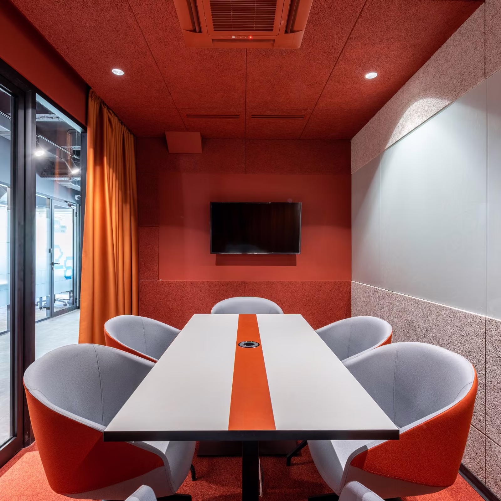 Modern acoustic meeting room with orange wall panels, a white conference table, grey chairs, wall-mounted screen, and glass partition.