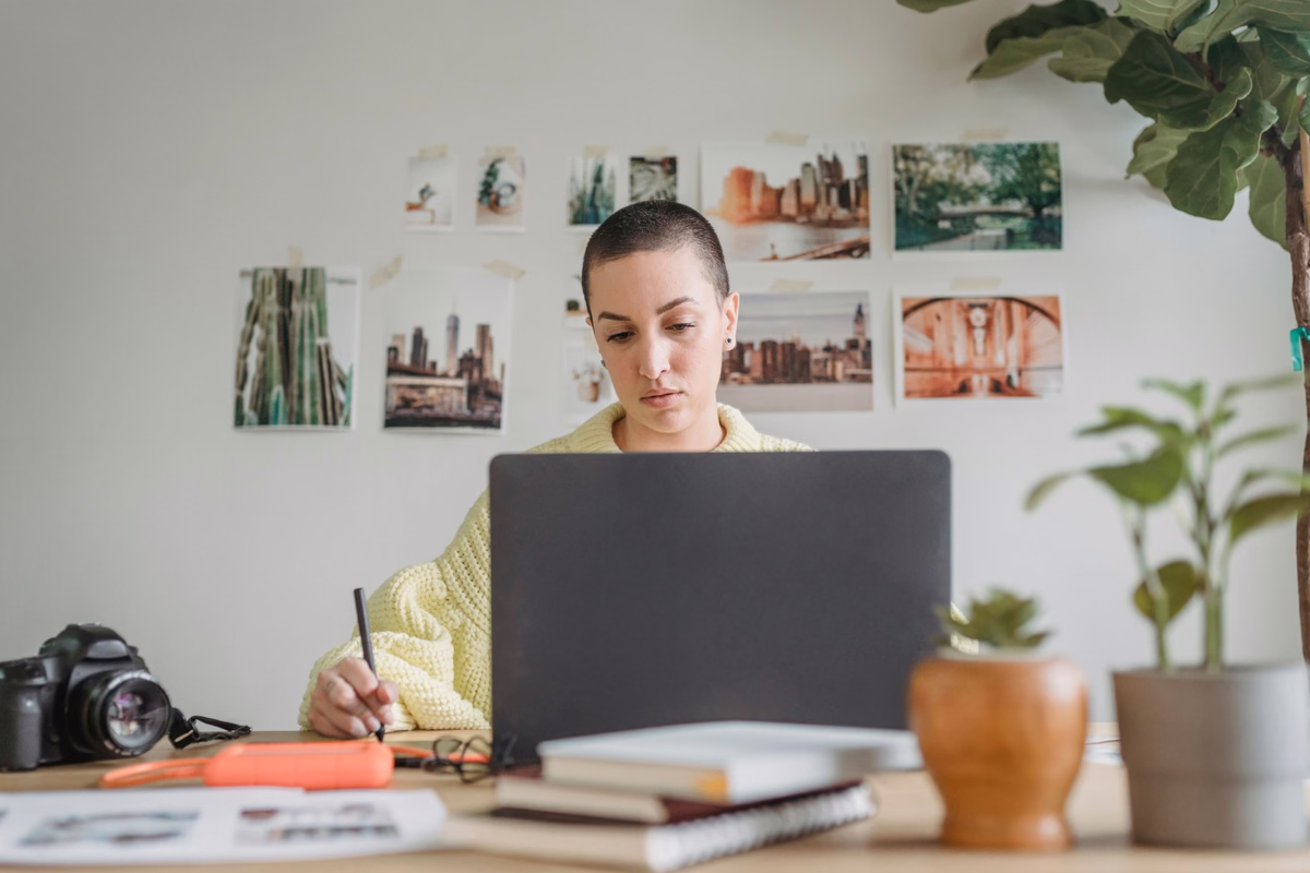 Woman working on a laptop and taking notes at a creative home-office desk, with a camera, notebooks, plants, and photos on the wall.