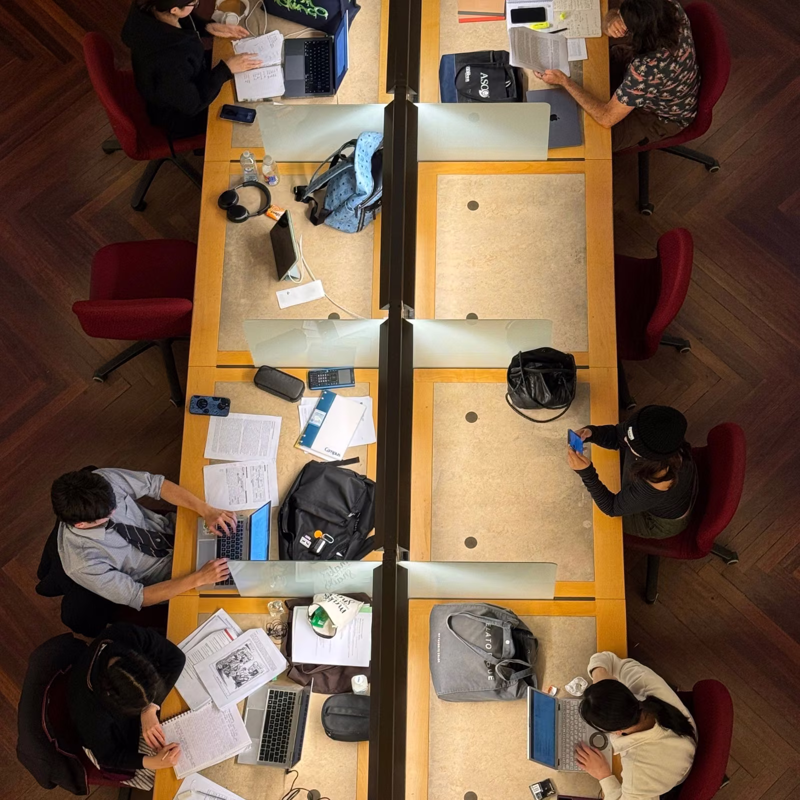 Overhead view of several people studying and working on laptops around a large shared table in a library or coworking space.