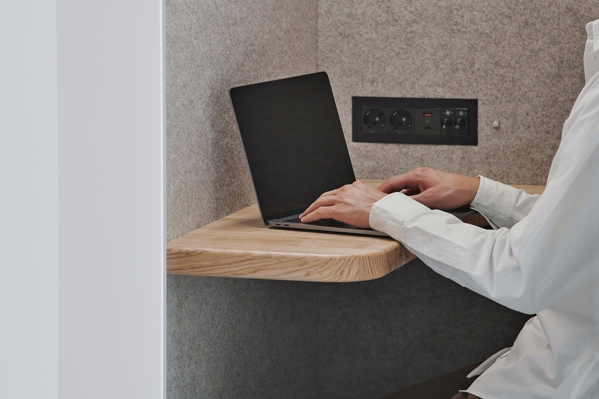 Person working on a laptop inside an acoustic office booth, with a wooden desk and integrated power outlets.