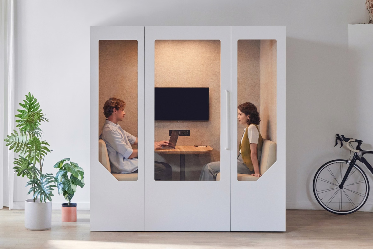 White acoustic meeting booth with two people seated inside, featuring a table, wall-mounted screen, warm felt interior, and glass panels, with plants and a bicycle in the surrounding office.