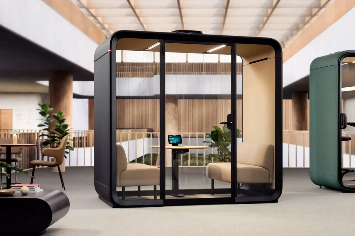 Modern black four-person acoustic meeting booth with glass walls, seating and a central table in a bright open-plan office.