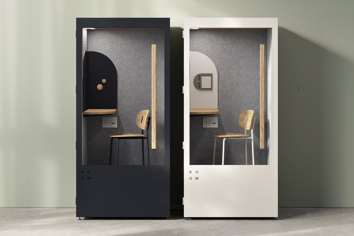 Two freestanding phone booths side by side against a sage-green wall: one in dark charcoal and one in cream, each with a grey acoustic felt interior, an oak fold-out shelf, a wooden chair, a power socket and a vertical oak handle.