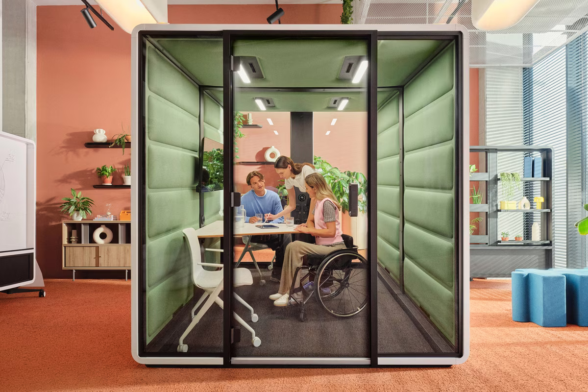 Glass-fronted office meeting booth with green acoustic panels, in an open-plan office with terracotta walls and carpet. Inside, three colleagues gather around a small table with a tablet.