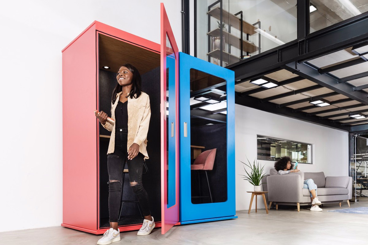 Two-section office phone booth, red on the left and blue on the right, in a bright loft-style workspace. A smiling woman steps out of the open red door holding a notebook, while another person relaxes on a sofa in the background.