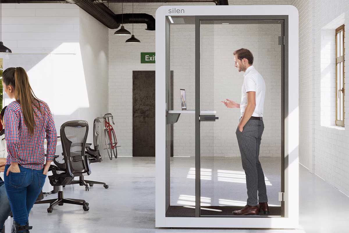 White phone booth with a glass door in a bright, industrial-style open office with white brick walls. A man in a white shirt stands inside talking, with a laptop on a small shelf behind him, while a woman walks past outside.