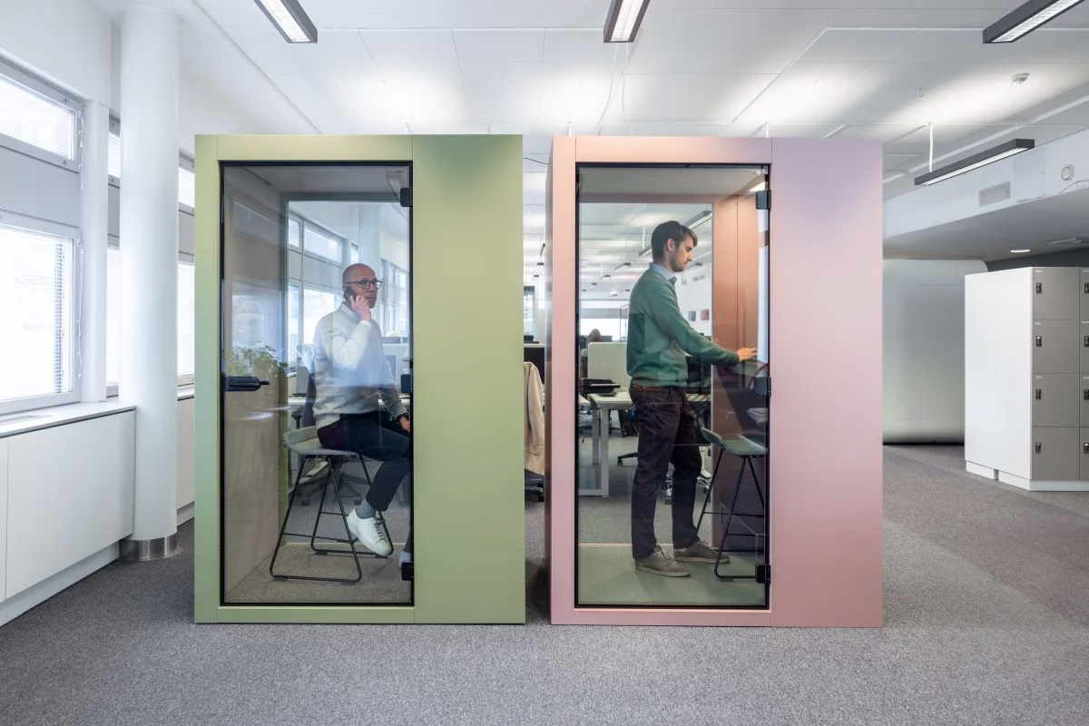 Two glass-fronted office phone booths side by side in a bright open-plan office, one with green panels and one with dusty pink panels. A man on a phone call sits on a stool in the green booth, and another man stands working at a shelf in the pink booth.