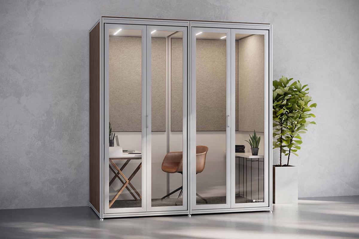Glass-fronted acoustic office booth with a desk and tan chair inside, beside a potted plant on a grey wall.