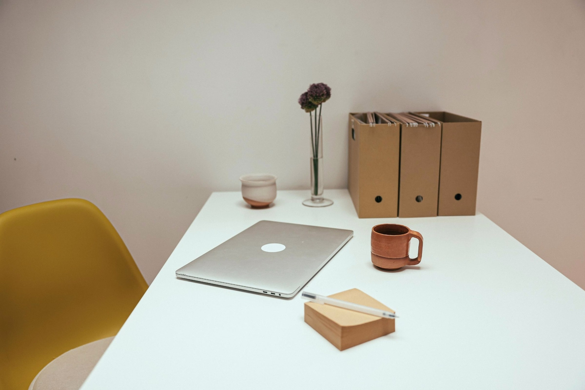 Minimalist workspace with a laptop, coffee mug, notebook and yellow chair at a white desk.