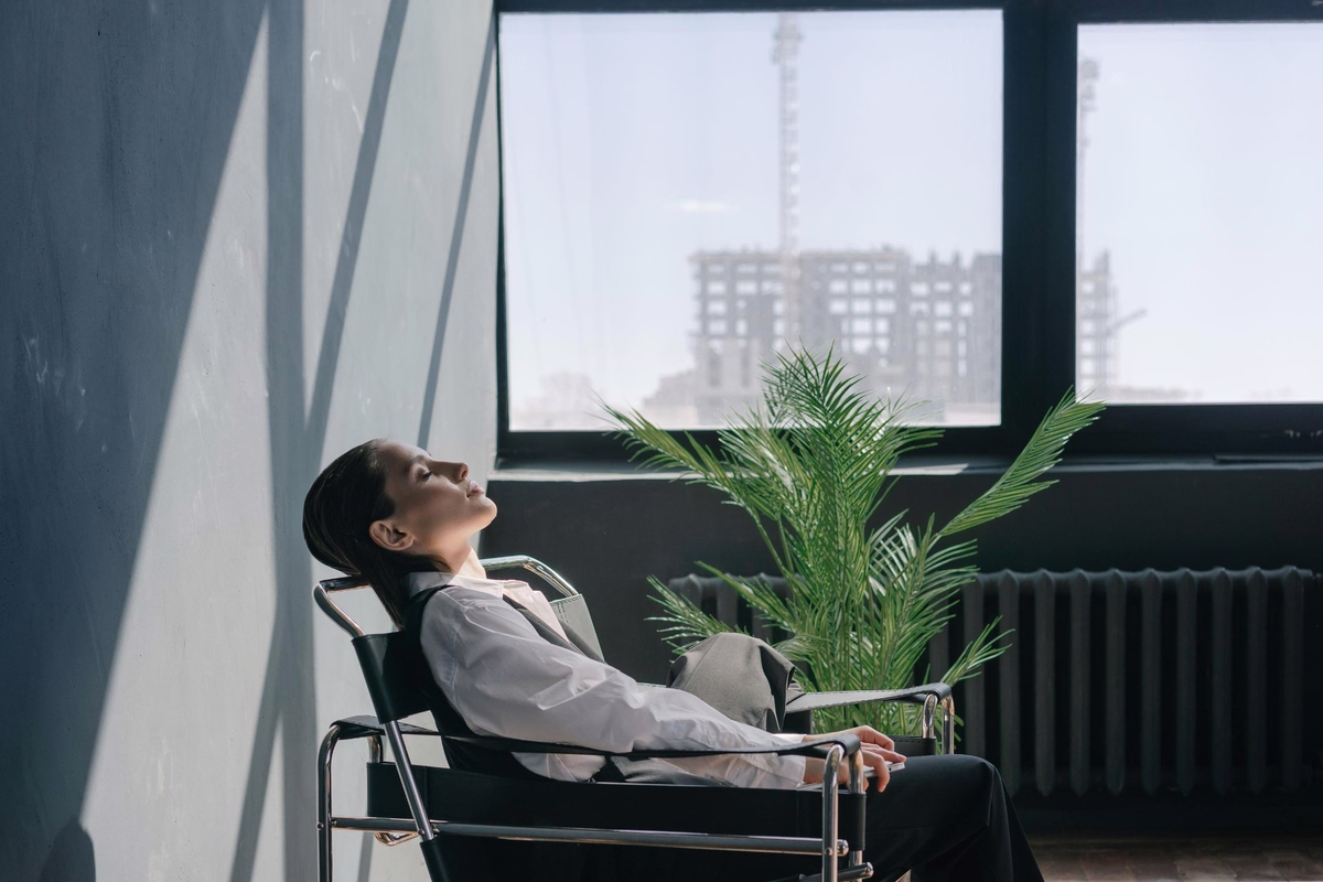 Professional woman relaxing in an office chair beside a sunlit window and indoor plant, enjoying a peaceful break at work.