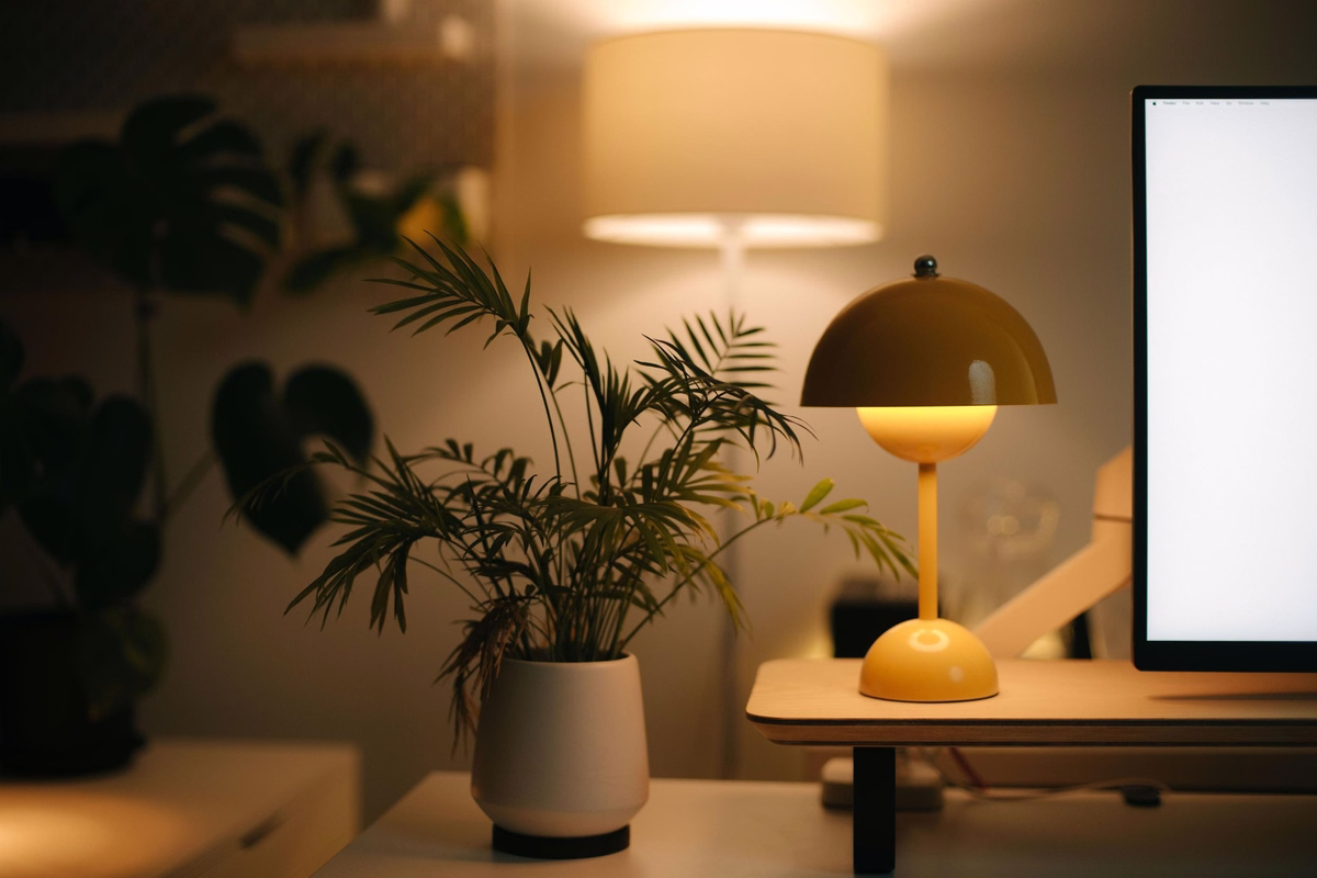 Cozy modern office workspace with warm ambient lighting, a yellow desk lamp, indoor plants and a computer monitor.