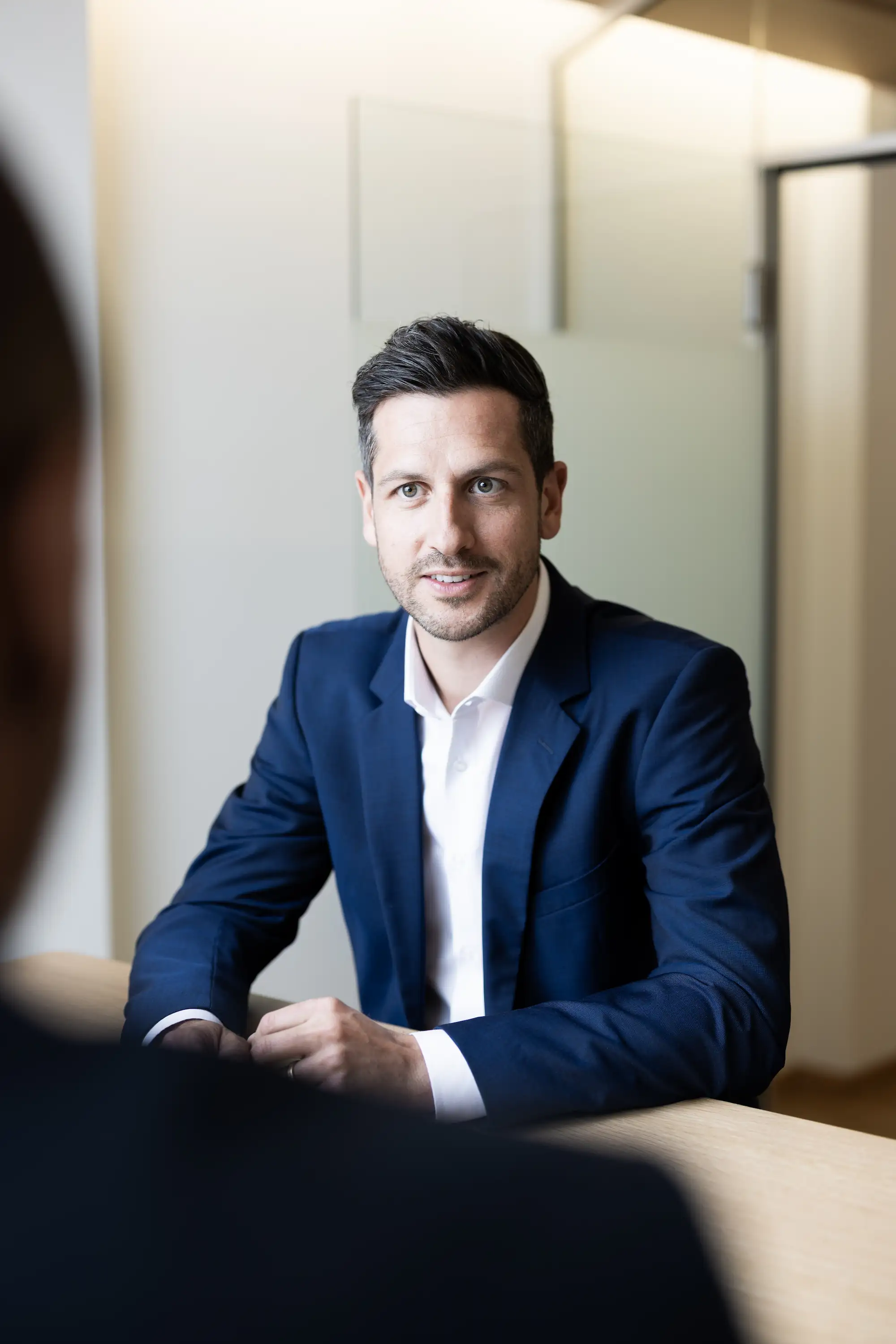 Yves Süess, Gründungspartner, in einem blauen Anzug und weißem Hemd sitzt an einem Tisch und spricht mit einer Person gegenüber.