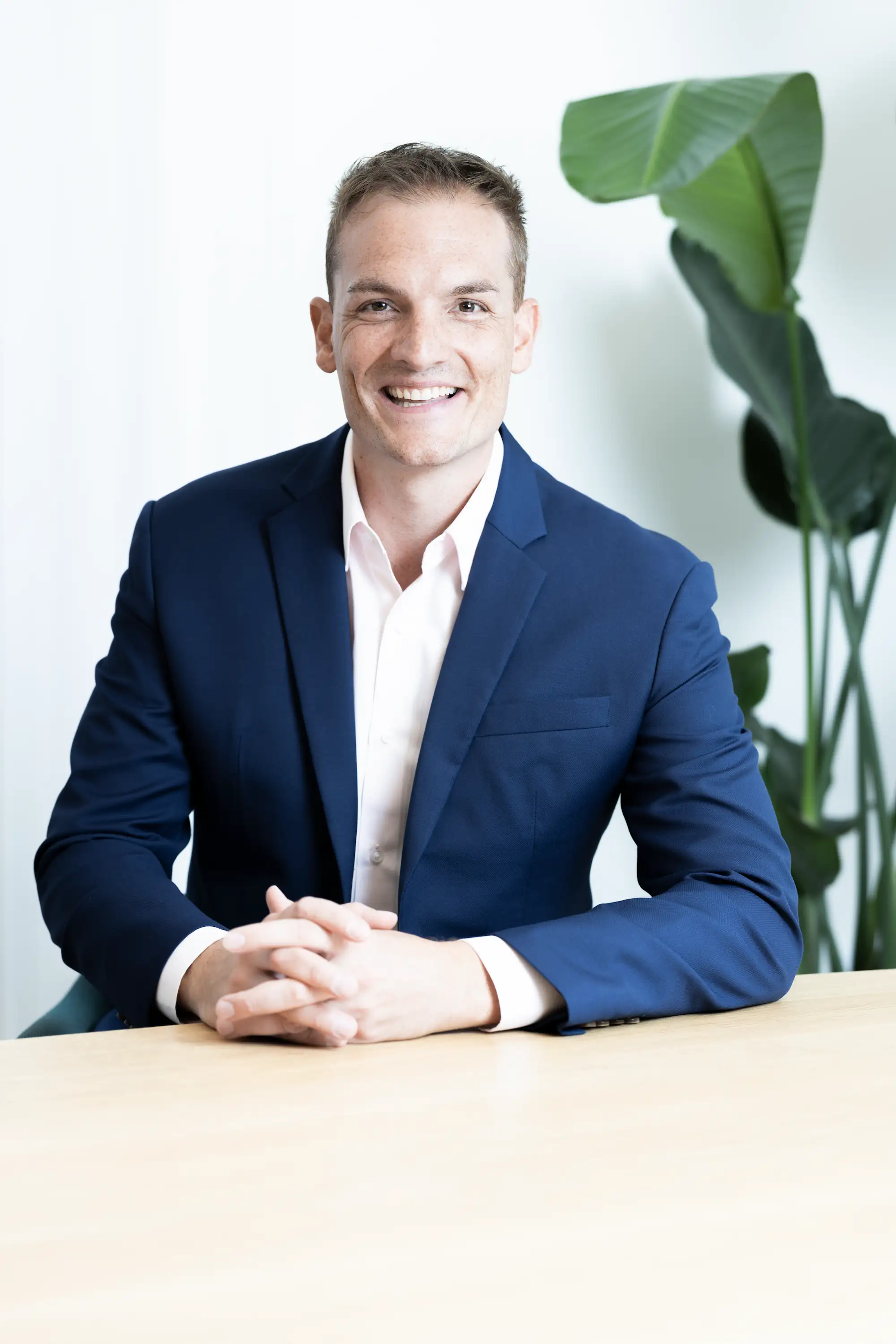 Fabian Rudin, Gründungspartner, lächelnd in blauem Anzug, sitzt mit verschränkten Händen an einem Tisch vor einem weißen Hintergrund und einer großen grünen Pflanze.