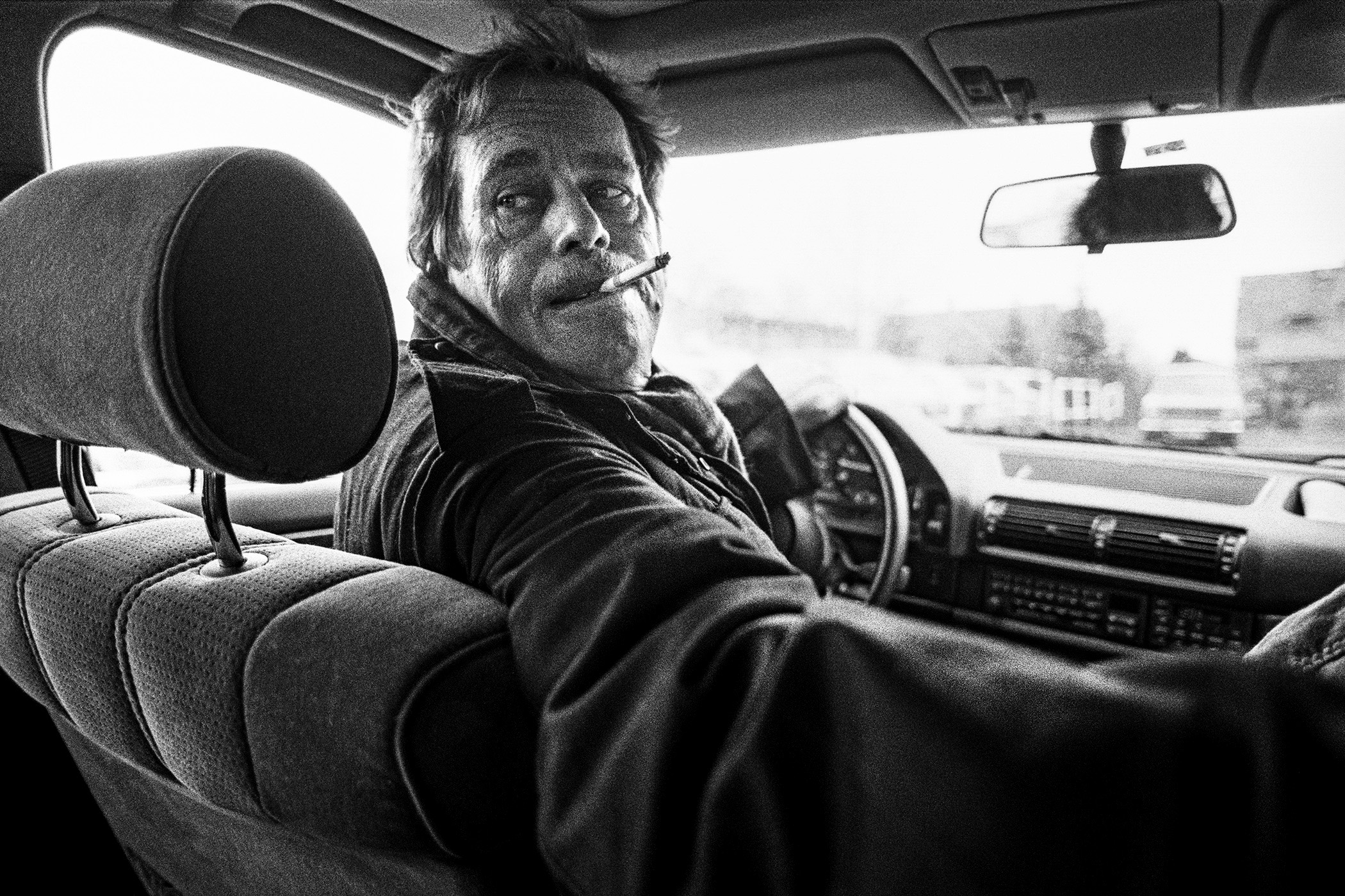 Prezident Václav Havel sedící za volantem auta, otočený dozadu s cigaretou v puse a výrazem gangstera.