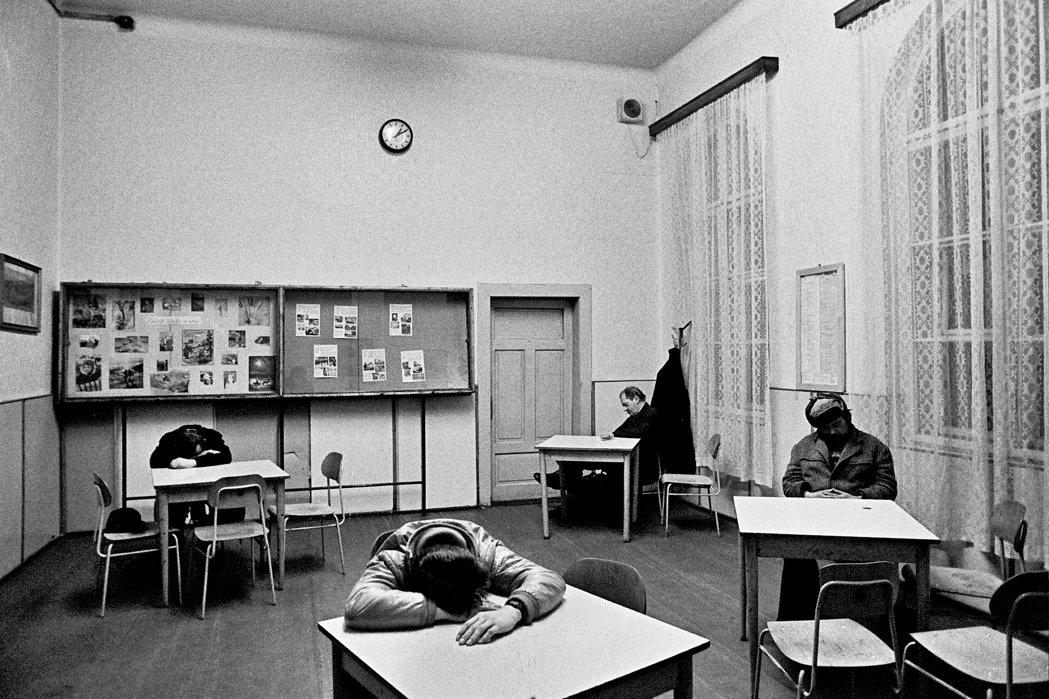 Skupina mužů u stolů spí v potemnělé nádražní hale v pozdních hodinách.