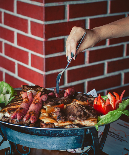 Una bandeja de metal llena de una variedad de carnes a la parrilla, salchichas y quesos, adornada con lechuga y tomates en rodajas a los lados.