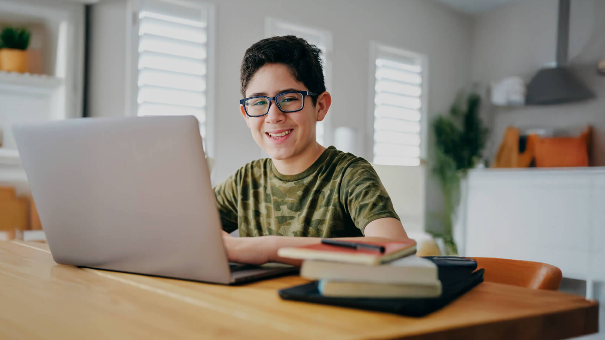 Boy wearing glasses smiles while using a laptop at a desk with books in a bright room.