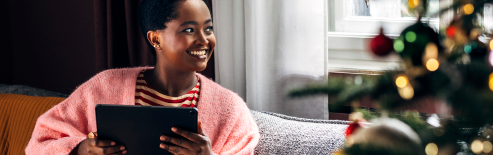 Smiling woman in a pink sweater holding a tablet and looking towards a decorated Christmas tree by a window.