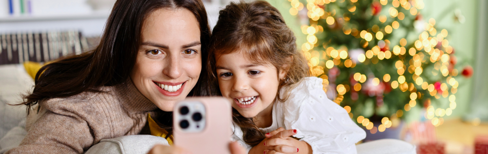 Smiling woman and young girl using a smartphone to make a video call in front of a decorated Christmas tree.