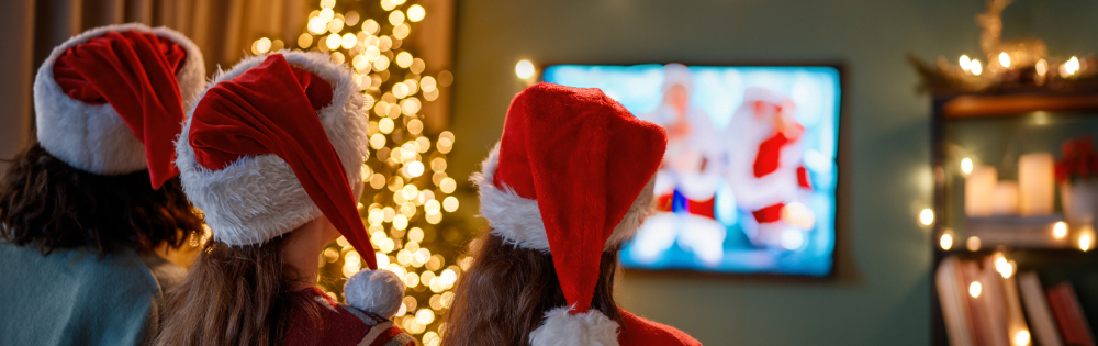 Three people wearing Santa hats watching a decorated Christmas tree and a blurred TV screen.