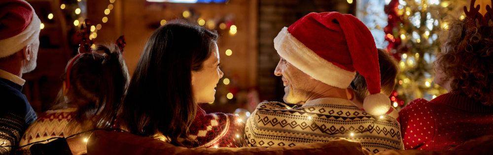 Group of people in festive sweaters and Santa hats sitting closely on a couch with warm holiday lights in the background.