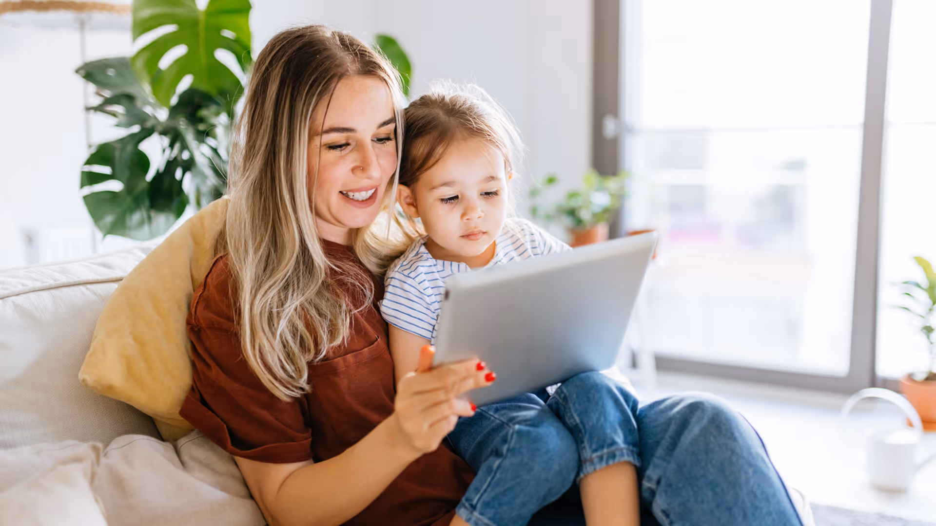 Woman and young girl sitting on a couch looking at a tablet together in a bright living room.
